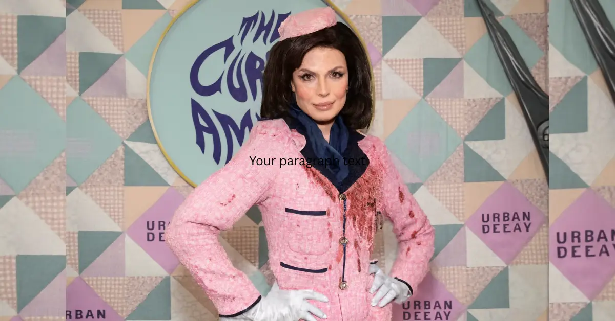 Julia Fox poses in a vintage-inspired pink tweed outfit with white gloves and a navy scarf at a Halloween-themed Urban Decay event, standing in front of a pastel geometric backdrop with oversized scissors.